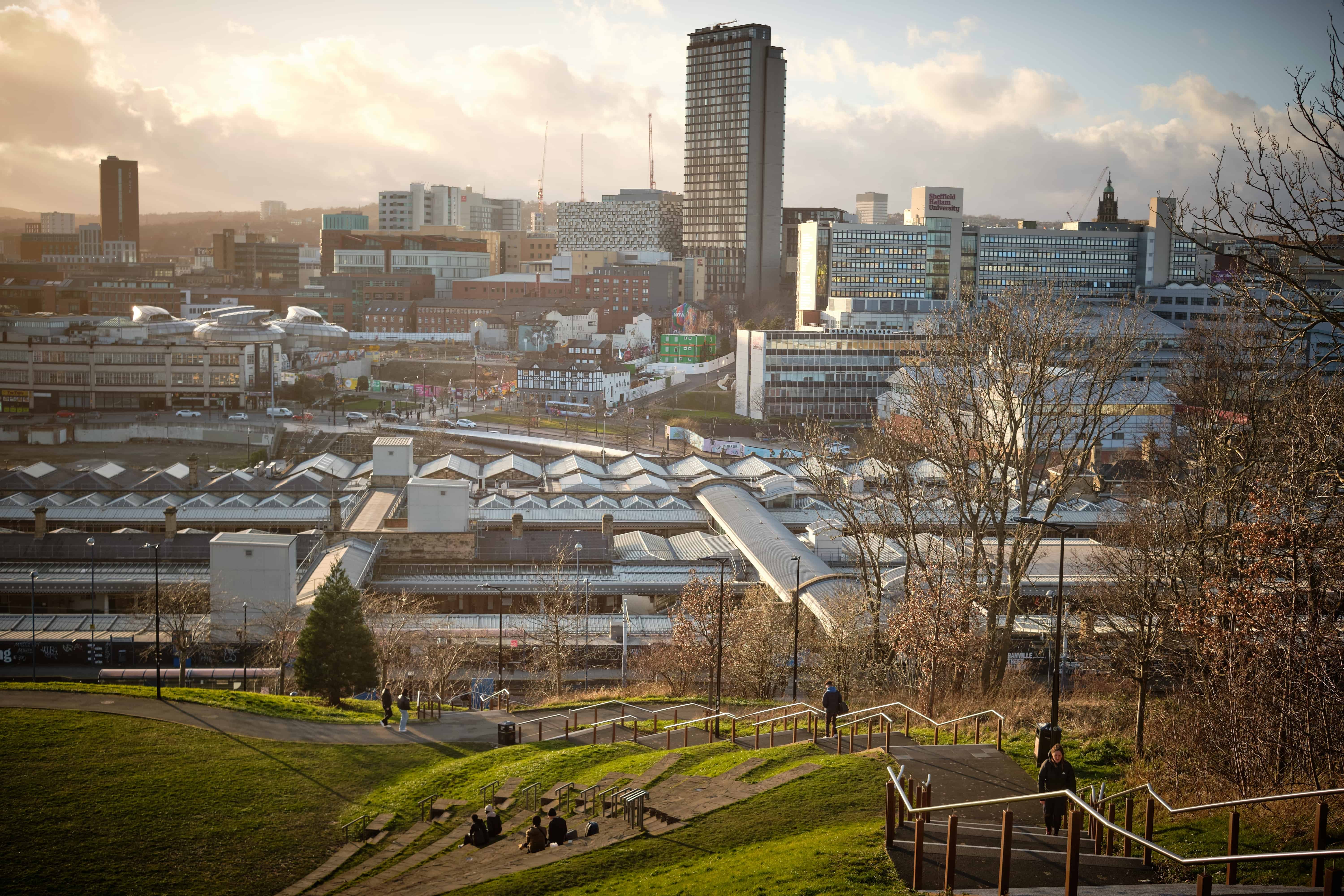 Sheffield city skyline with modern buildings, Sheffield Hallam University, and train station rooftops viewed from a hillside park, highlighting the city’s growth and connectivity.